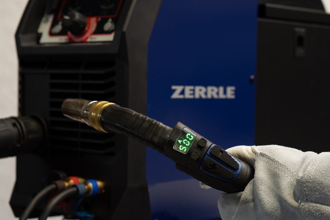 Hand im Schweißerhandschuh hält einen MIG/MAG-Brenner mit Digitaldisplay vor einem Zerrle-Schweißgerät